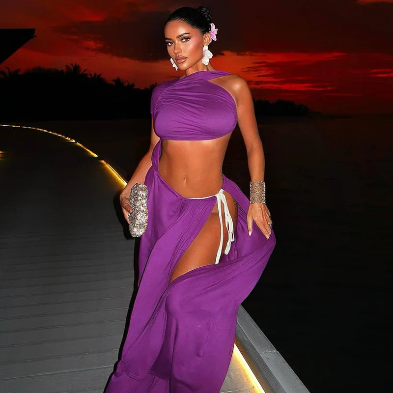 A woman models a vibrant purple two-piece lounge set consisting of a halter crop top and a matching high-slit wrap skirt, accessorized with a sparkly silver clutch and statement earrings, set against a sunset background.
