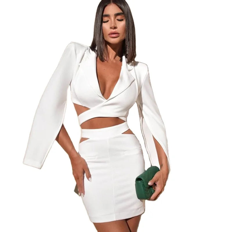 A woman modeling a white two-piece outfit consisting of a long-sleeve wrap-style blazer with cutouts and a matching high-waisted mini skirt, holding a small green clutch against a plain white background. Baby boo nadine merabi clubL London