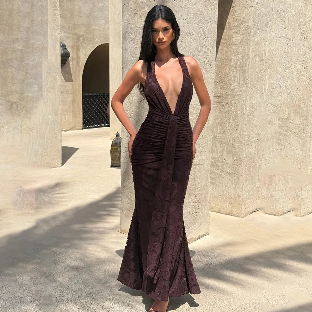 A woman stands outdoors in a sunlit architectural setting wearing a sleeveless, deep V-neck, ruched mermaid gown in a dark plum-burgundy tone. The dress has a textured jacquard-like pattern and a flared hem. babyboo ClubL NAdine merabi Bronx and banco elisabetta revolve