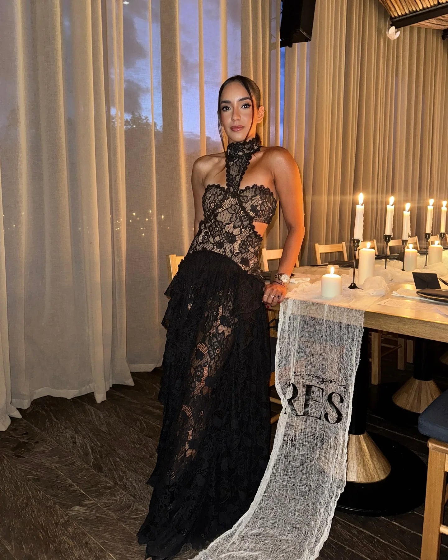 Woman in a black lace dress standing in a restaurant with 'RES' on a chair.